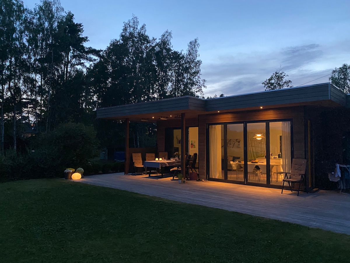 A modern cabin is illuminated softly in the evening, showcasing large glass doors that provide a view into a well-lit interior. Outdoor seating areas are arranged on a spacious wooden deck, surrounded by a lush green lawn and trees in the background.