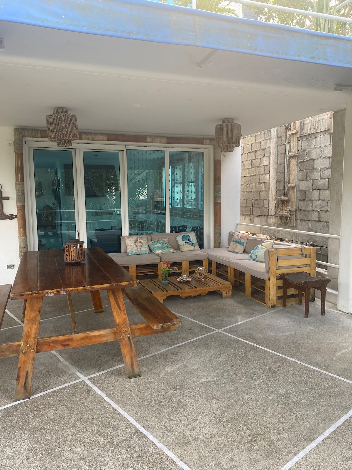 An outdoor seating area features a wooden table alongside a comfortable bench adorned with decorative pillows. Chairs are positioned nearby, while large glass doors provide a view of the interior. Natural light illuminates the space, highlighting the simplicity and comfort of the setting.