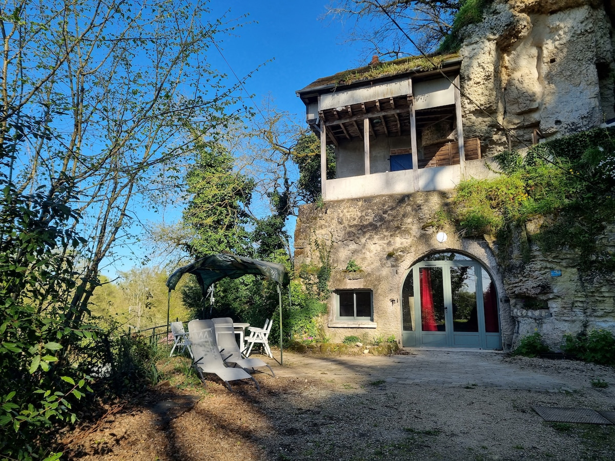 The exterior of the troglodyte house is showcased, with a terrace featuring two reclining lounge chairs under a shaded canopy. Lush greenery and trees surround the area, creating a serene environment. The entrance is visible, framed by natural stone and clear skies.