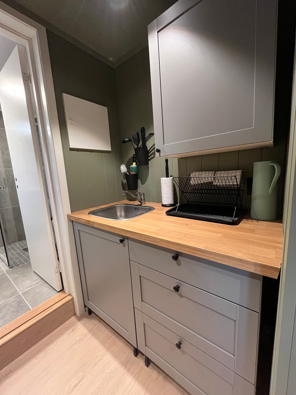 A compact kitchenette is displayed with a wood countertop and grey cabinetry. A small sink is positioned next to a drying rack, with storage drawers below. Green walls create a soothing backdrop, and a doorway visible leads to a tiled bathroom area.