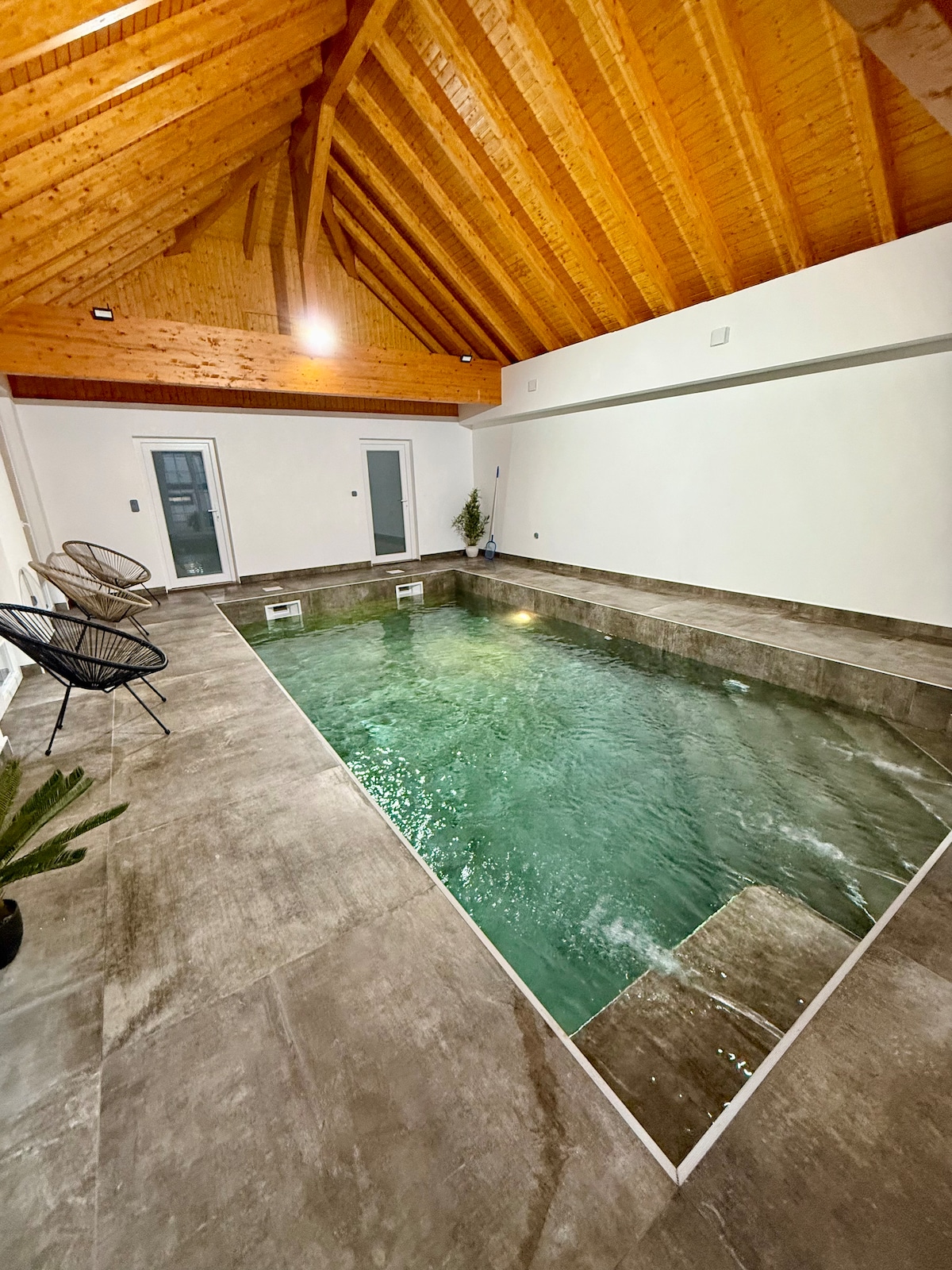 An indoor swimming pool with a calming green hue is surrounded by sleek stone flooring. The space features a high wooden ceiling with exposed beams, enhancing the open feel. Two stylish black chairs are positioned nearby, creating a relaxing atmosphere for unwinding.