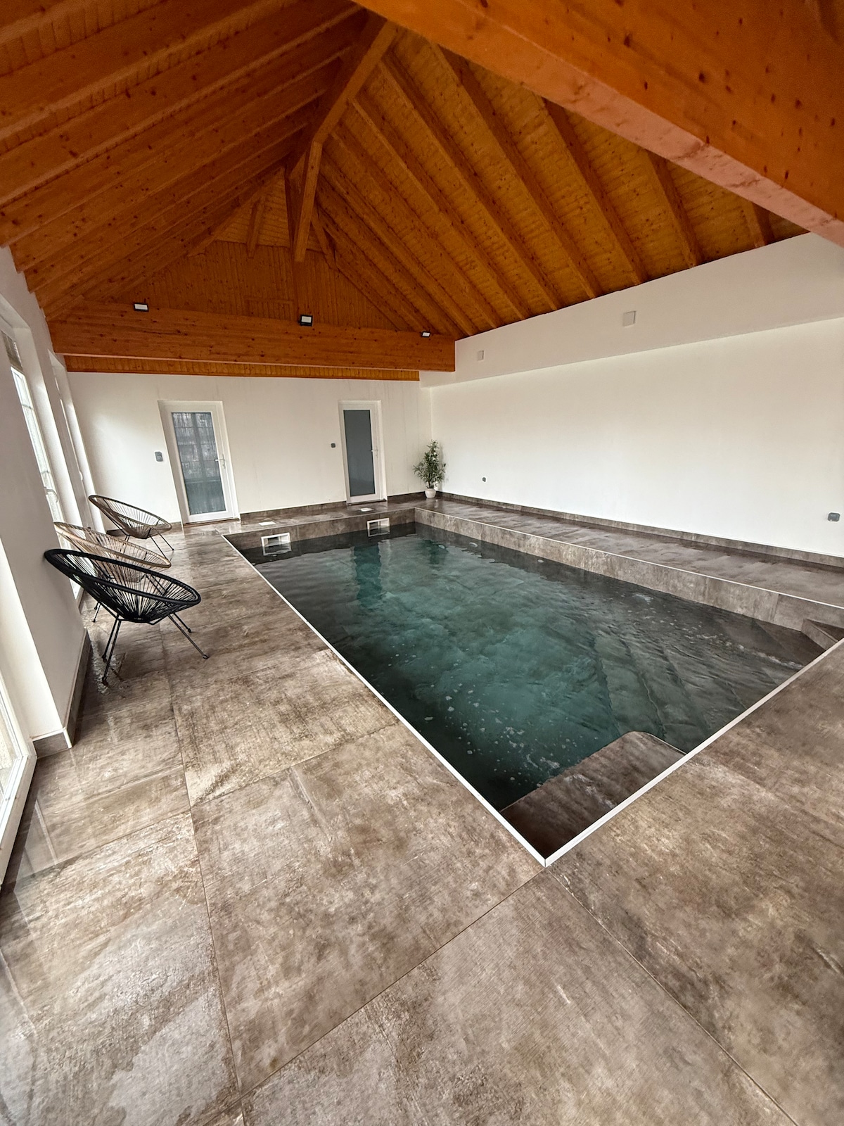 An indoor heated pool is showcased within a spacious area, featuring light-colored walls and a wooden ceiling that highlights the design. Two black chairs are positioned near the poolside, adding to the serene ambiance.