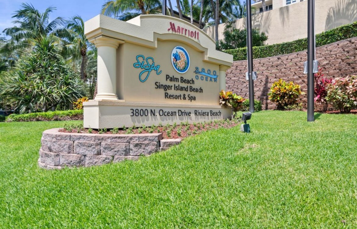 A welcoming entrance sign for the Palm Beach Marriott® Singer Island Beach Resort & Spa is displayed prominently amidst lush green landscaping. Tropical plants and palm trees surround the sign, conveying a sense of relaxation and inviting guests to the resort.