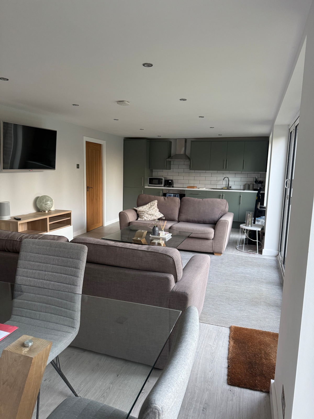 A spacious living area is visible, featuring a comfortable grey sofa set facing a flat-screen TV mounted on the wall. A modern kitchen with green cabinetry is seen in the background, alongside a dining table with stylish grey chairs, creating an inviting communal space.