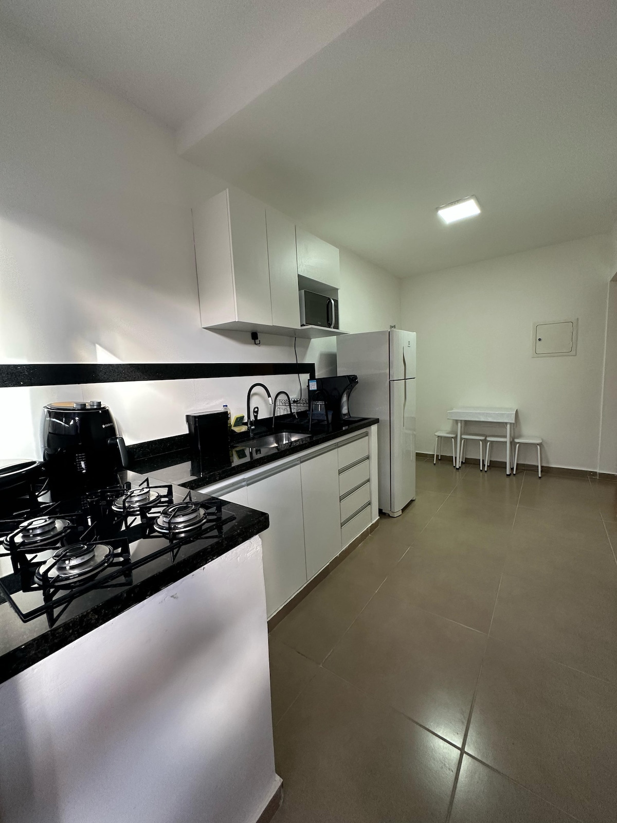 The kitchen area is equipped with sleek white cabinetry and black granite countertops. Modern appliances include a stove with five burners, a microwave, and a coffee maker. A small dining table with two chairs is positioned in the corner, under bright ceiling lighting.