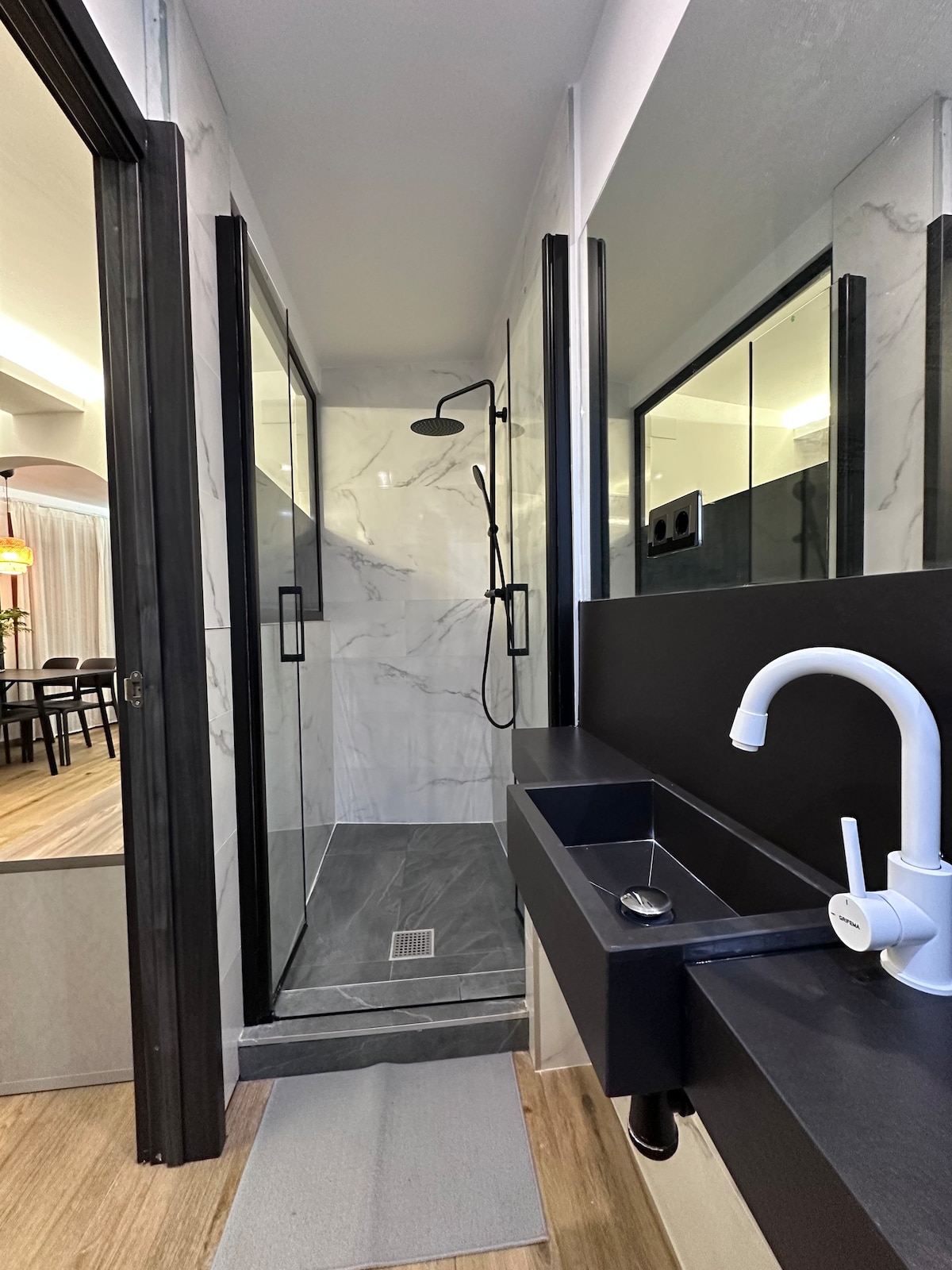 A modern bathroom is highlighted with sleek marble walls and a spacious walk-in shower. The shower features a rainfall showerhead mounted above, complemented by glass doors. A stylish black sink and faucet add to the contemporary design, with a soft mat placed on the floor.