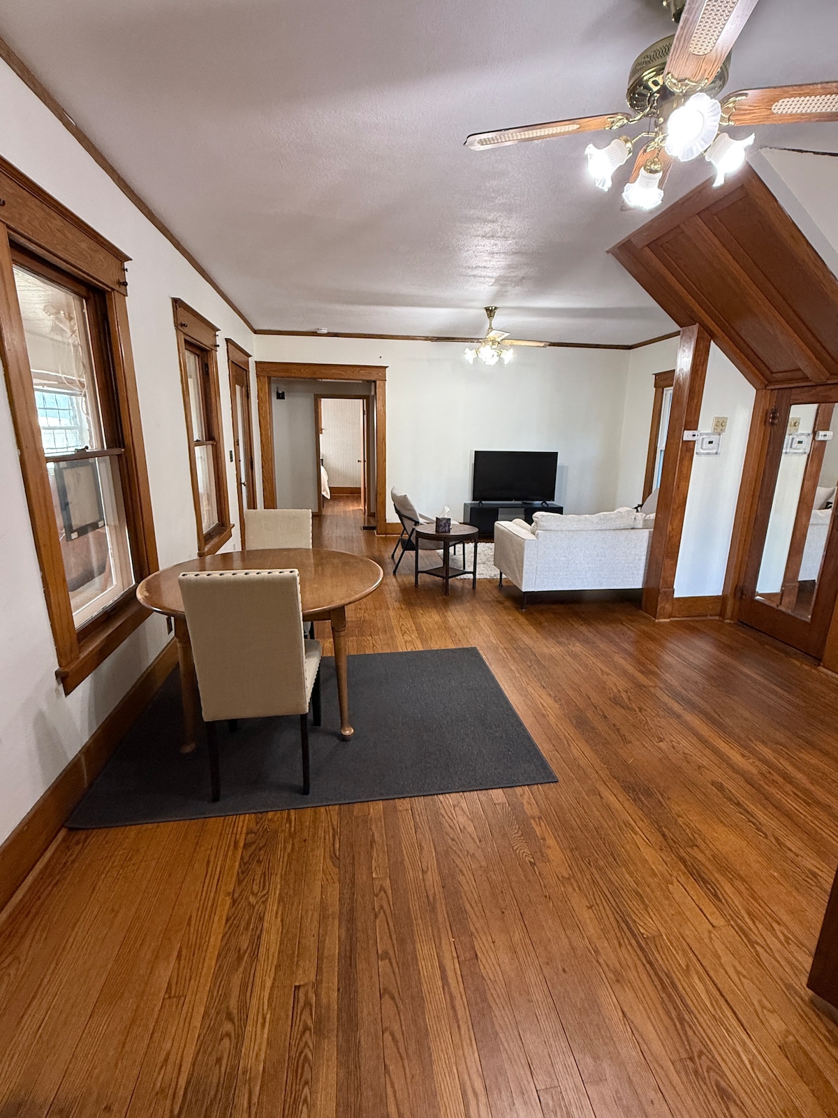 A welcoming living area showcases polished wooden floors and rich wood trim. A round dining table with two chairs sits adjacent to a cozy seating arrangement. A television is mounted on the wall, and natural light fills the space through multiple windows.
