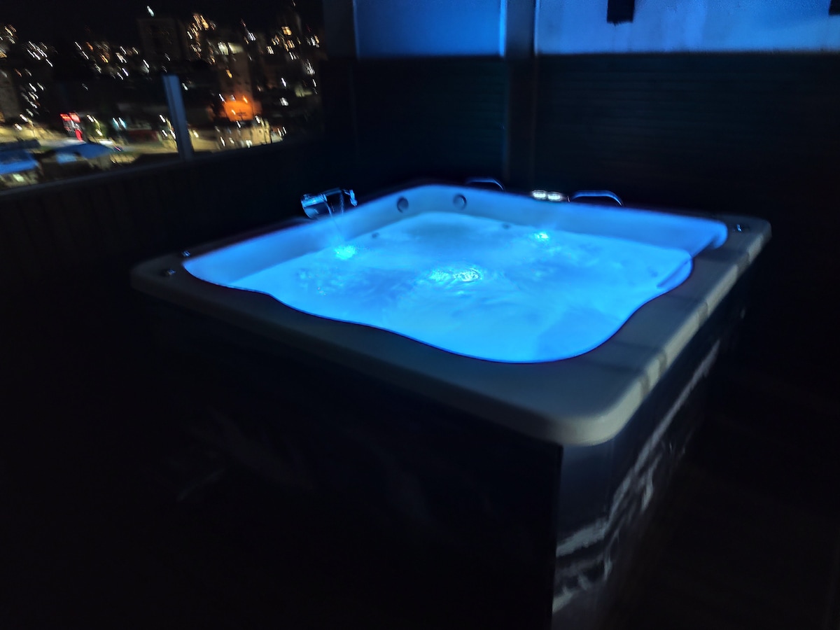 A hot tub is illuminated with soothing blue lights, creating a relaxing atmosphere. The tub is situated on a wooden deck with city lights visible in the background, enhancing the tranquil setting for guests to unwind.