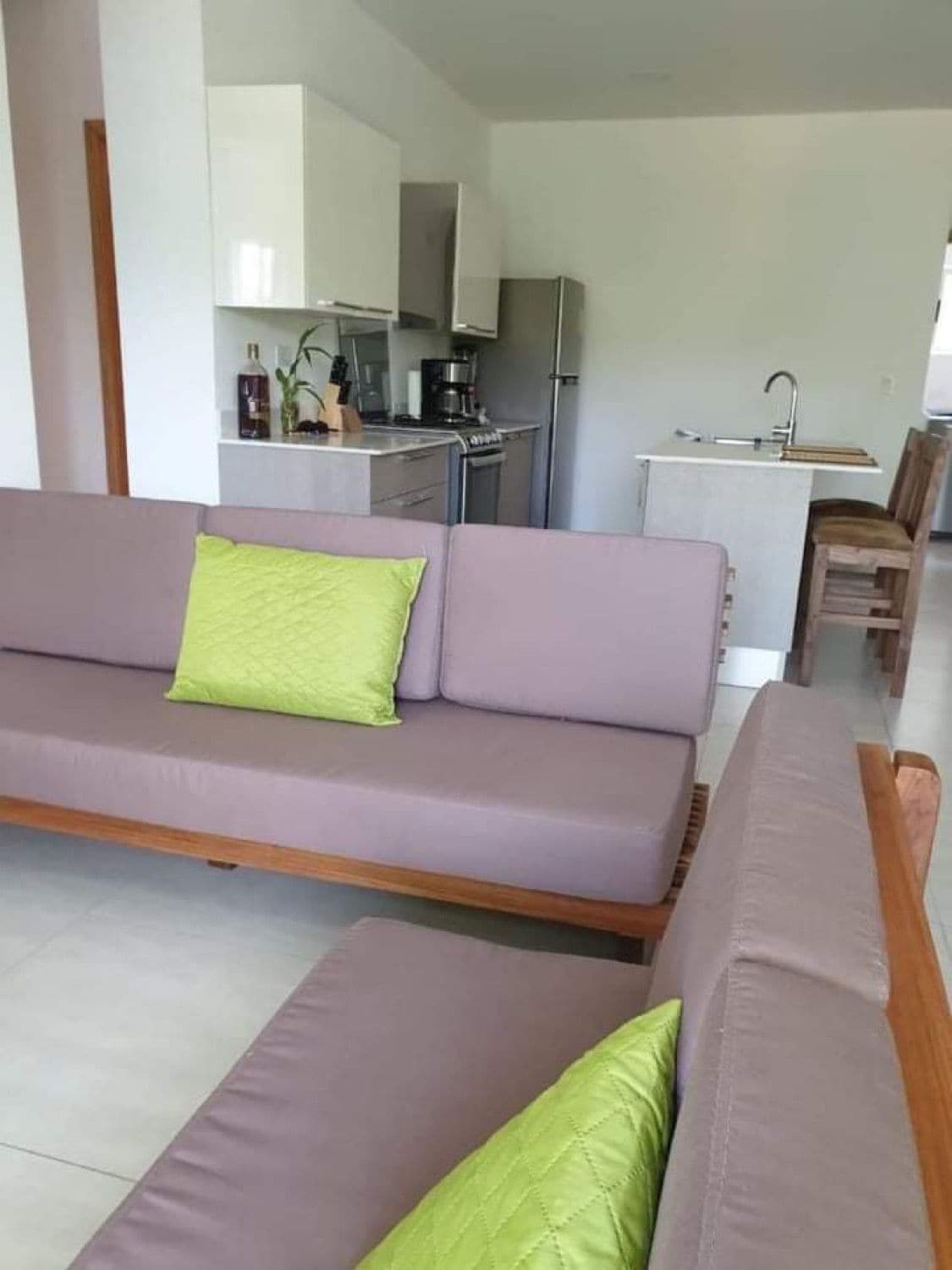An inviting living area features a pair of wooden sofas adorned with lime green cushions. In the background, a modern kitchen with stainless steel appliances is visible, showcasing an open layout with bar seating at the counter.