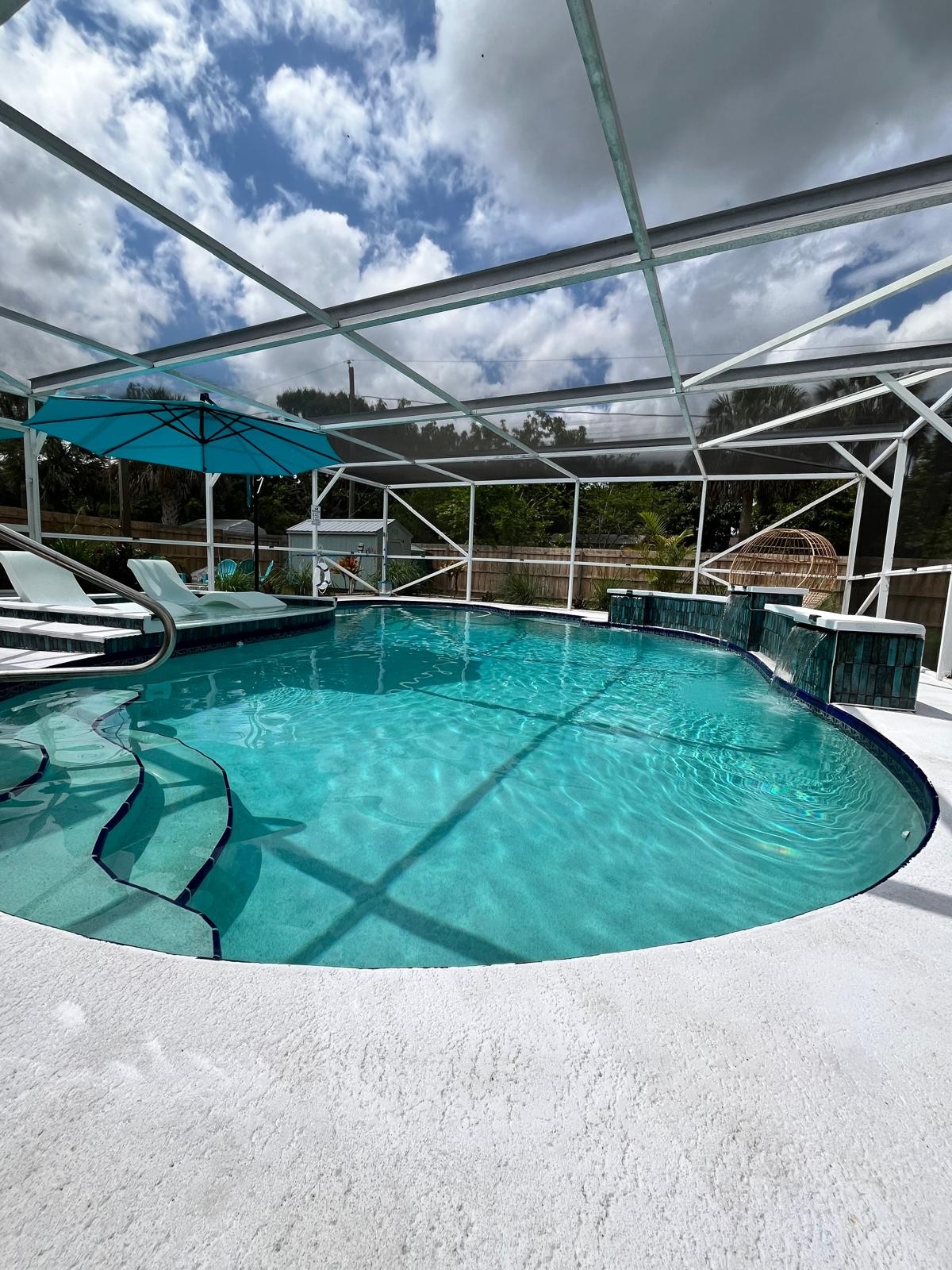 An outdoor pool area is enclosed by a screened patio, featuring a circular pool highlighted by clear blue water. Poolside loungers are positioned under an umbrella, providing shade. Natural light filters through the mesh panels, creating a serene environment with lush greenery visible in the background.