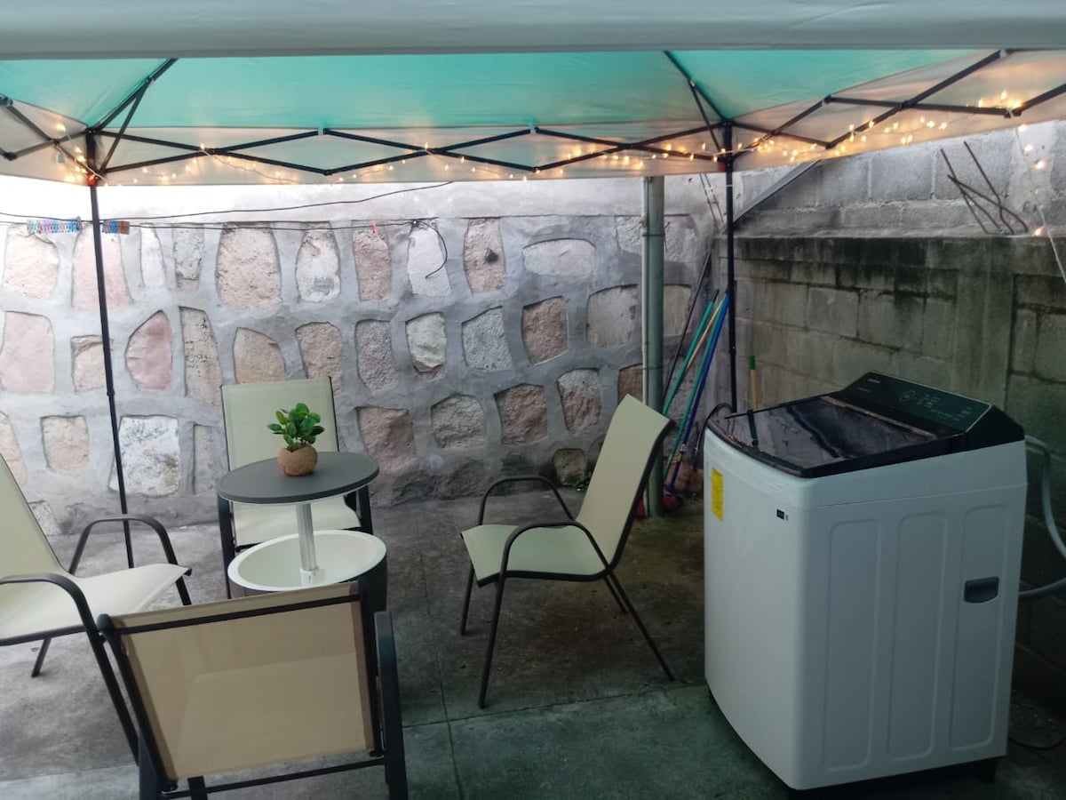 A cozy outdoor space is covered by a tent. Underneath, a small table is surrounded by two chairs, and a potted plant adds a natural touch. A washing machine is positioned against a stone wall, with string lights softly illuminating the area.