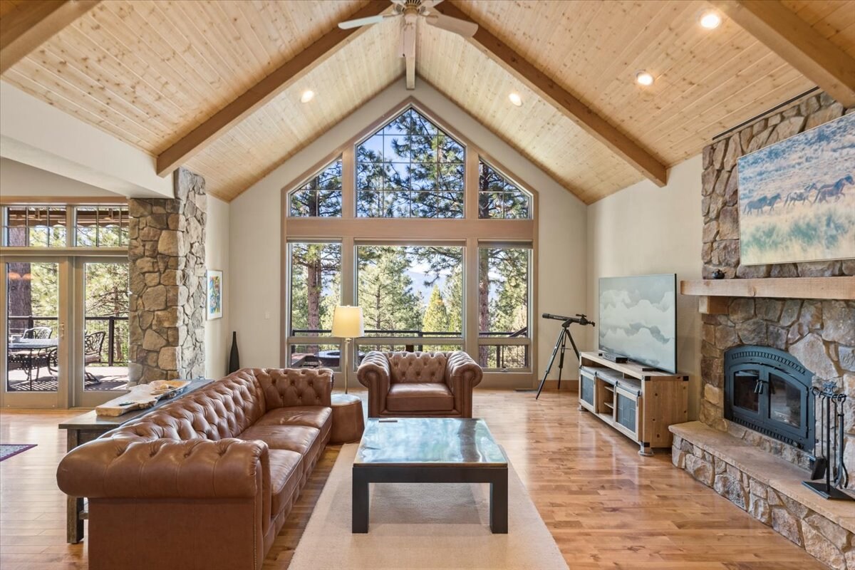 A spacious living area features high wooden ceilings and large windows allowing for ample natural light. A cozy leather sectional and a pair of classic armchairs surround a wooden coffee table. A stone fireplace adds a rustic touch, and a telescope is positioned near the window, inviting exploration of the outdoors.