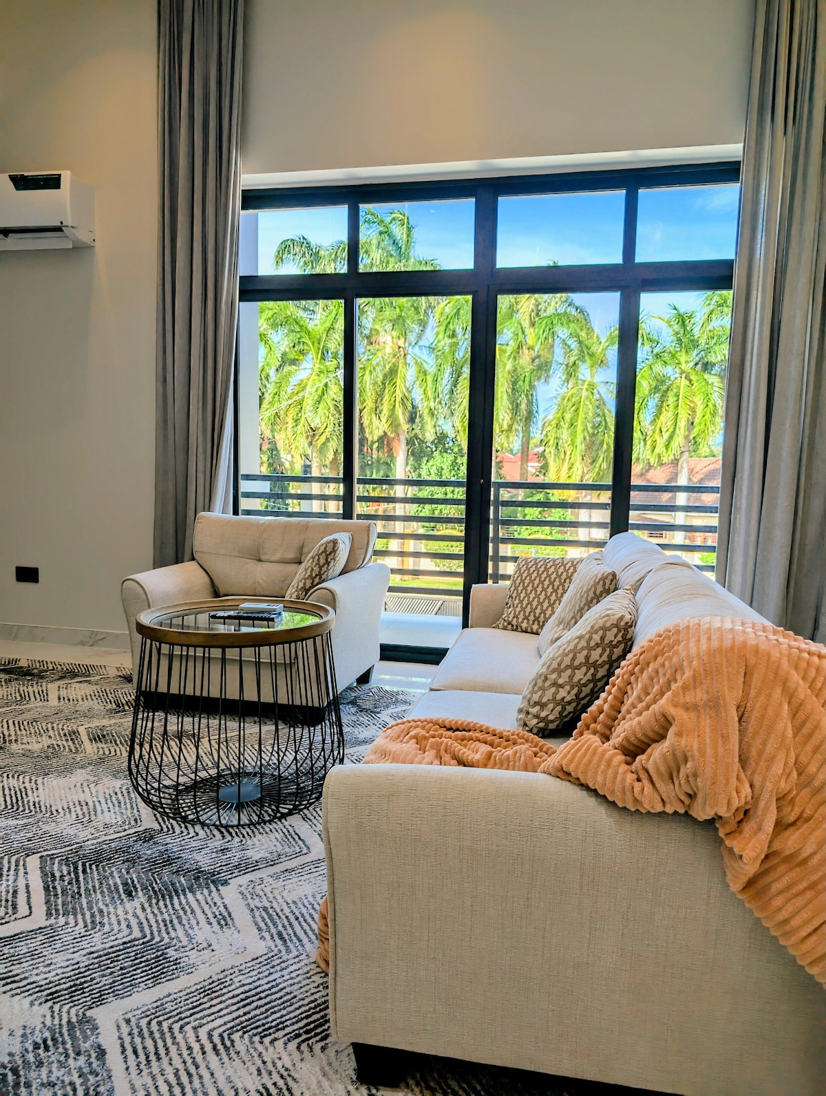 A light-filled sitting area features two comfortable chairs and a cozy sofa draped with a soft throw. A round wire coffee table sits in the center, with palm trees visible through the large windows, offering a fresh and serene outdoor view.