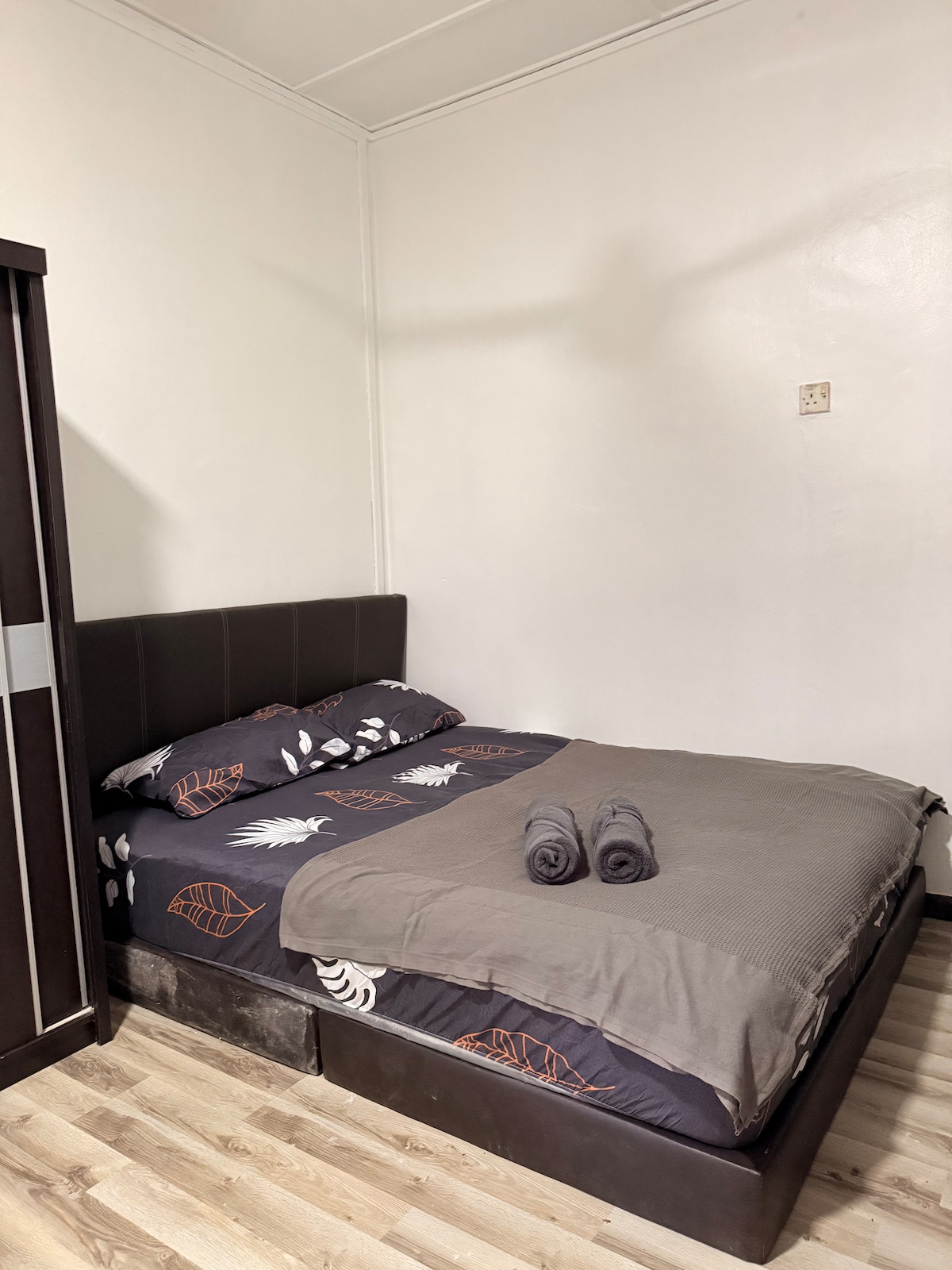 A neatly arranged sleeping area features a spacious bed with a dark, patterned duvet cover and two fresh towels placed at the foot. A dark wardrobe stands against one wall, and light-colored flooring completes the room's layout.