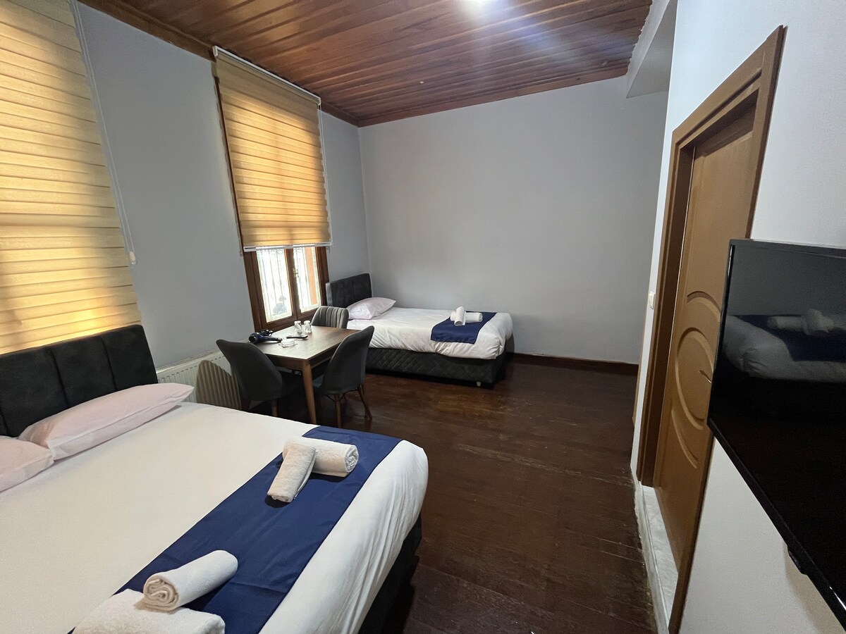 An airy family room is displayed with two spacious beds and a small dining table. Light-colored wooden flooring is present, complemented by gentle natural light filtering through the window blinds. A wall-mounted television adds to the amenities of the space.