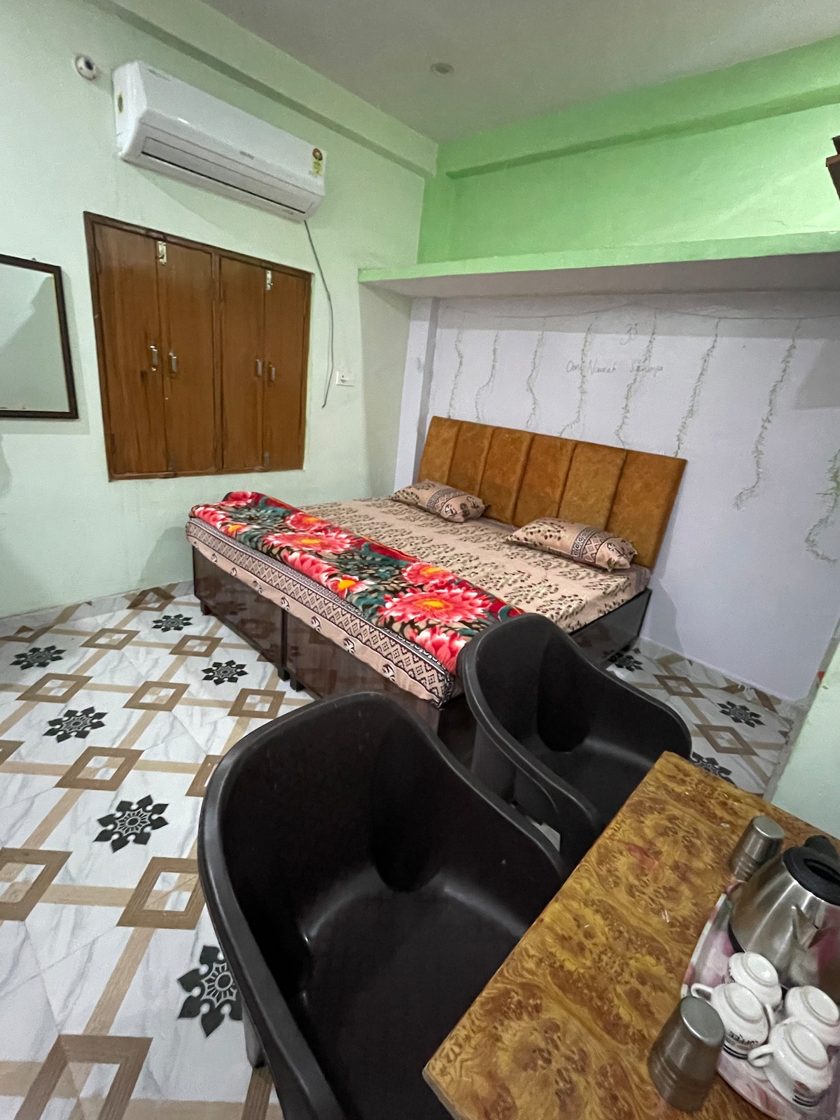 A cozy room is presented with a spacious bed adorned with vibrant floral bedding. Two cushioned chairs are positioned at a small table. Natural light enters through a window, and built-in cabinets are visible on one wall, providing storage options.