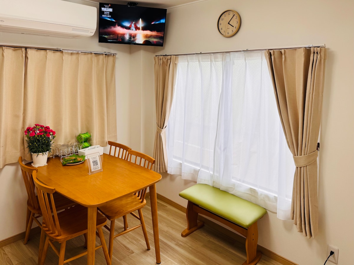 A dining area features a wooden table surrounded by four chairs, complemented by a green bench. Natural light filters through a window covered by sheer curtains. A small clock is mounted on the wall. A television is mounted above, showcasing a scenic image.