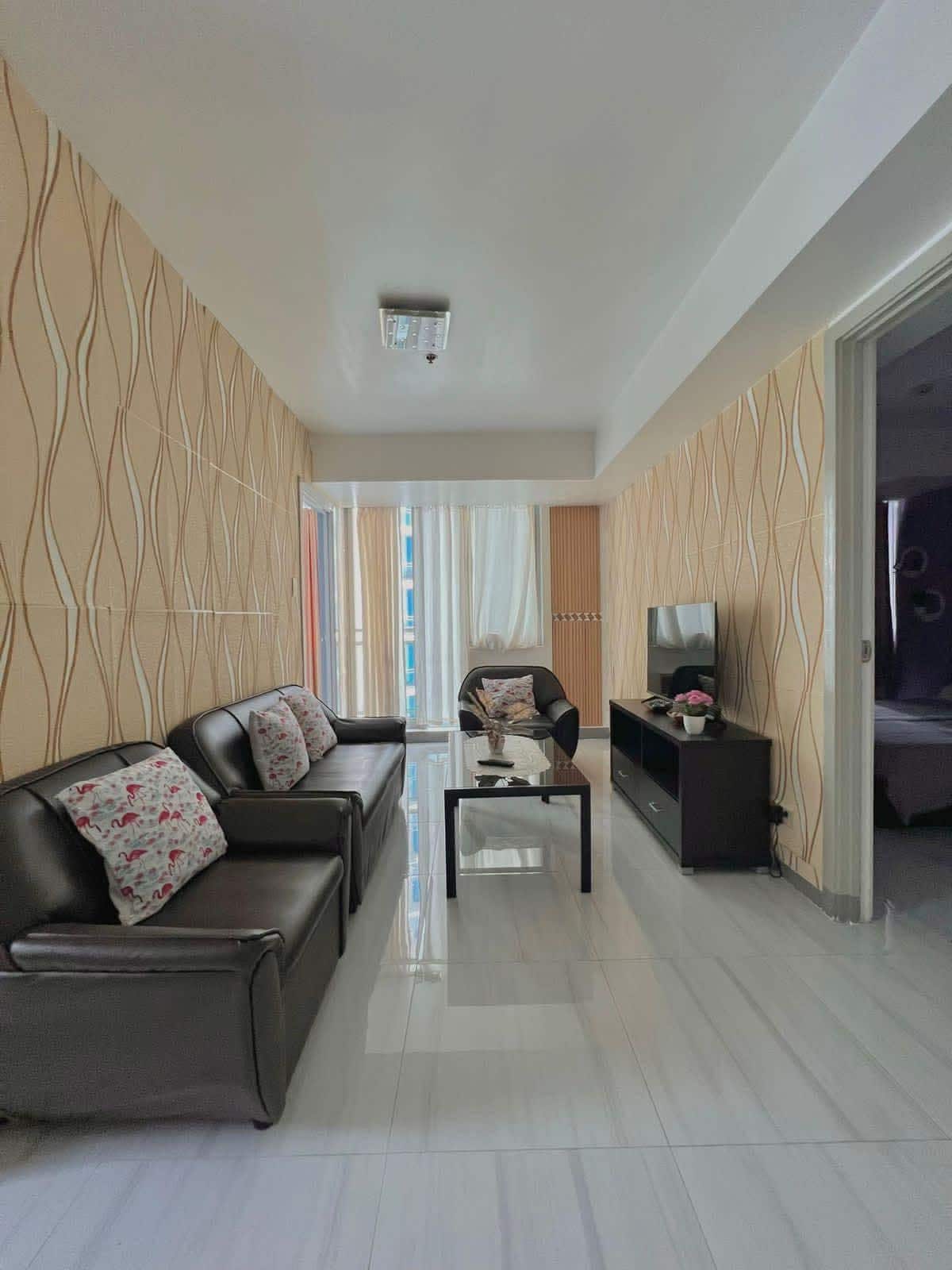 A living area is presented, featuring two dark leather sofas with floral throw pillows and a coffee table at the center. A television on a low stand completes the space, while large windows allow natural light to illuminate the room, creating a bright and open atmosphere.