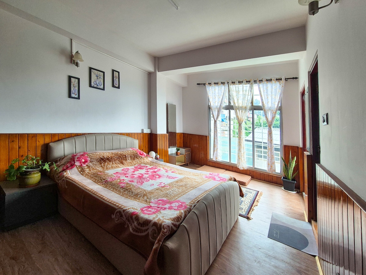 A spacious room features a large bed with a decorative blanket, positioned against a wooden wall. Natural light filters through large windows adorned with sheer curtains, illuminating a small indoor plant near the floor. Decorated walls include framed artwork, contributing to a calming environment.