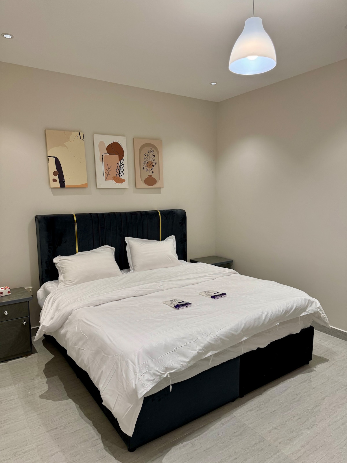 A modern bedroom displays a spacious bed with crisp white linens and two fluffy pillows. The dark upholstered headboard adds contrast to the light-colored walls. Above the bed, three art pieces are arranged, complementing the overall aesthetic. A bedside table is positioned on each side.