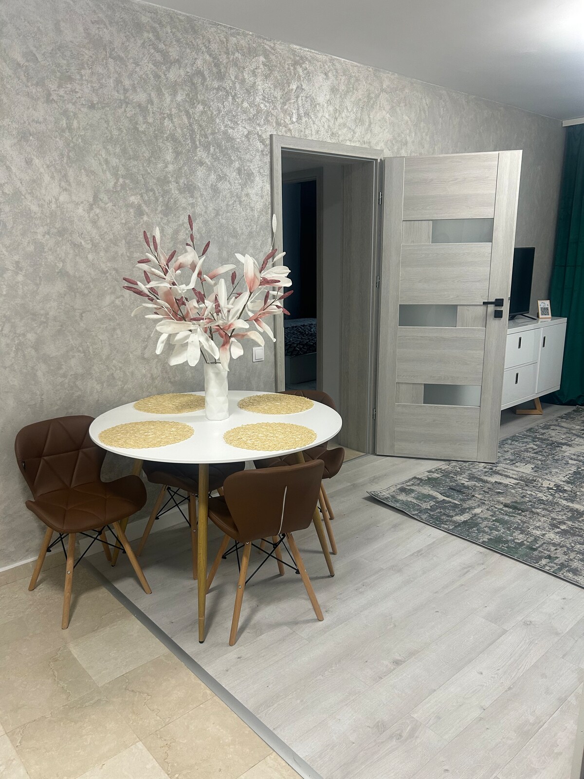 A round dining table with a white surface is surrounded by four brown chairs. A vase filled with decorative branches occupies the center of the table. A door to another room is visible in the background, contributing to the open layout of the space.