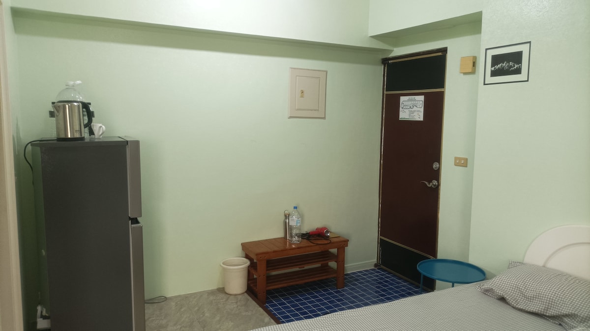 A room featuring light green walls and a large window providing natural light. A spacious bed is paired with a wooden table and chairs. A refrigerator and kettle are visible, along with a water bottle on the table. The entrance door is marked for easy identification.