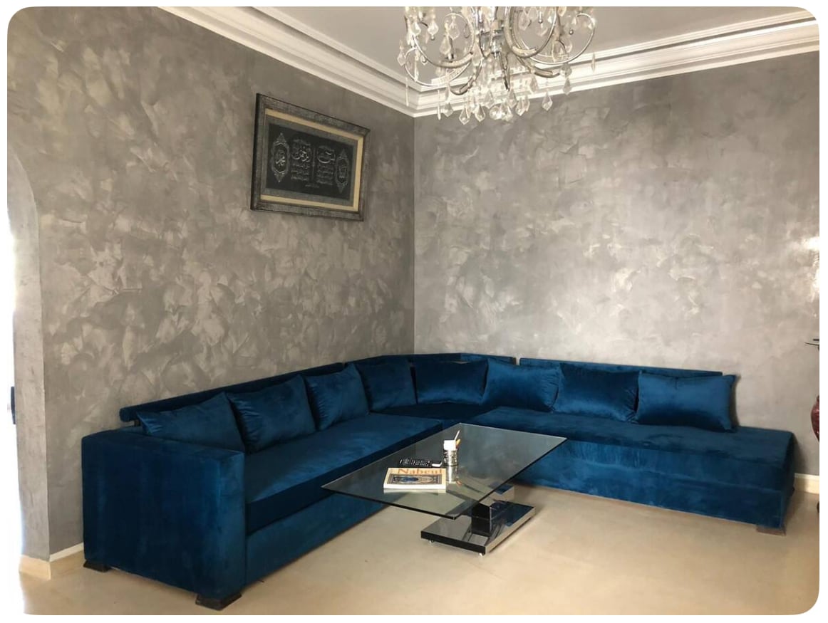 A spacious blue velvet sectional sofa is arranged around a glass coffee table. The textured gray wall adds depth, while a decorative piece hangs above the sofa. A modern chandelier provides overhead lighting in this inviting living area.