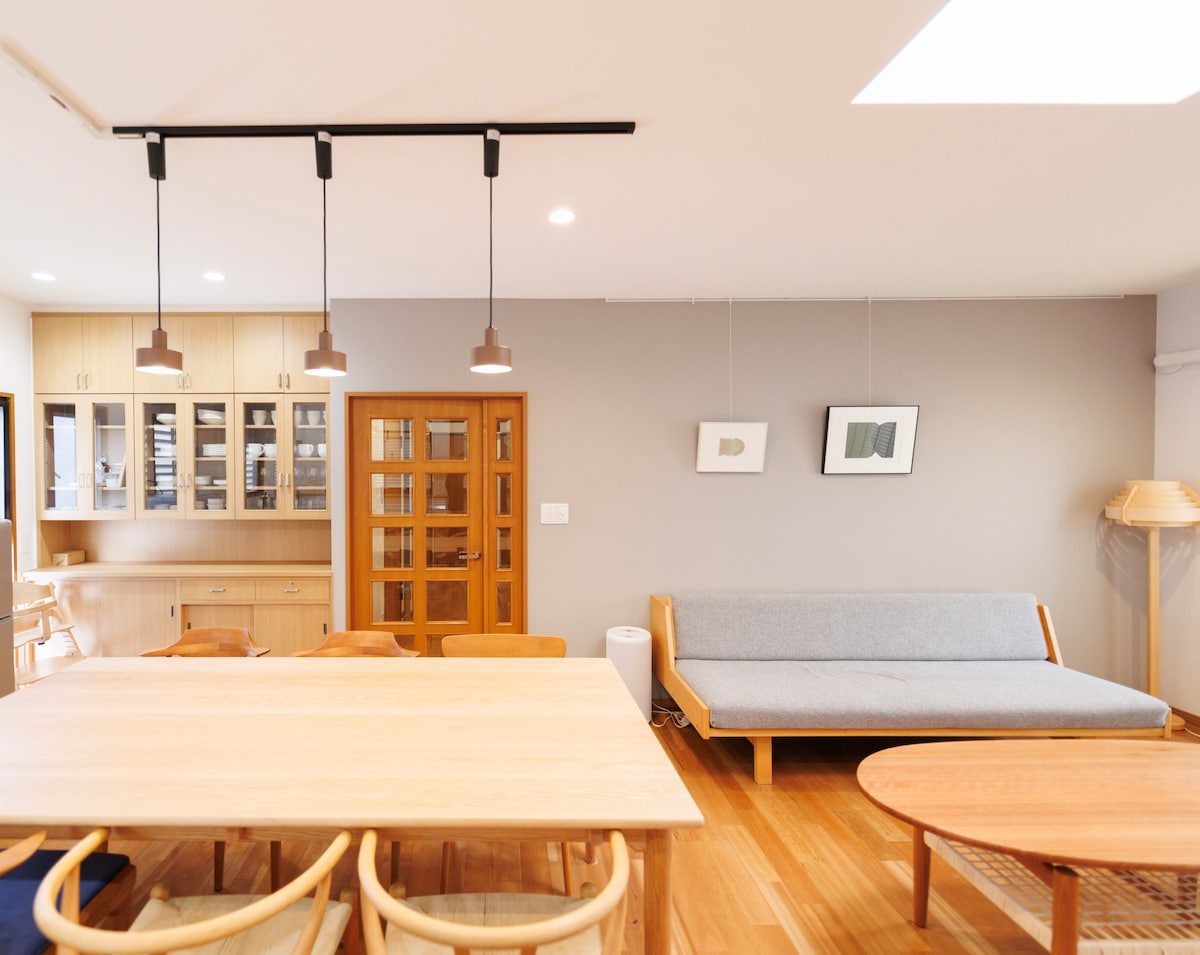 A spacious dining area features a long wooden table surrounded by light wood chairs. A minimalist sofa rests against the wall near artwork. Soft lighting is provided by pendant fixtures, illuminating the light wood flooring and contrasting gray walls.