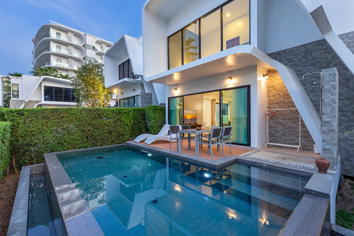 A private swimming pool is featured in the outdoor area, framed by lush greenery. The patio includes a dining set, providing a space for meals outside. Large glass doors lead into a modern living area, seamlessly connecting indoor and outdoor environments.