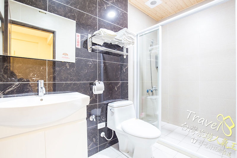 The bathroom features modern tile work with a walk-in shower enclosed by glass. A compact white sink and toilet are positioned against a dark marble wall. Shelves with neatly arranged towels are mounted above the sink, providing easy access for guests.