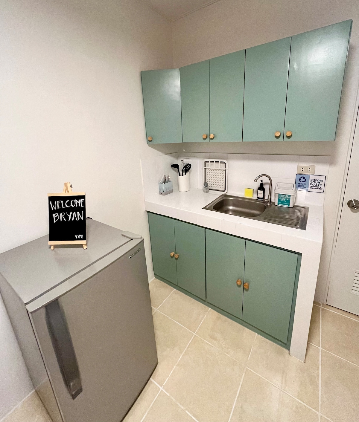The kitchenette features light green cabinets and a compact stainless steel refrigerator. A small sign welcomes guests, and a sink with necessary kitchen essentials is visible. Cooking utensils are organized neatly on the countertop, providing a functional space for light meal preparation.