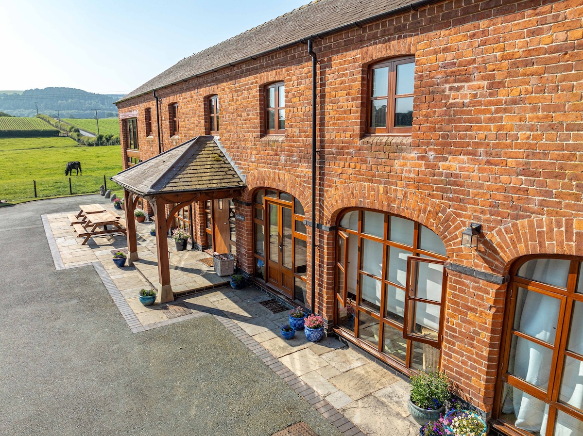 The exterior of a brick building features large arched windows, providing ample natural light. A sheltered entrance with a wooden roof offers a welcoming space for guests. Surrounding flower pots add a touch of color. The landscape beyond presents fields of greenery under a clear blue sky.