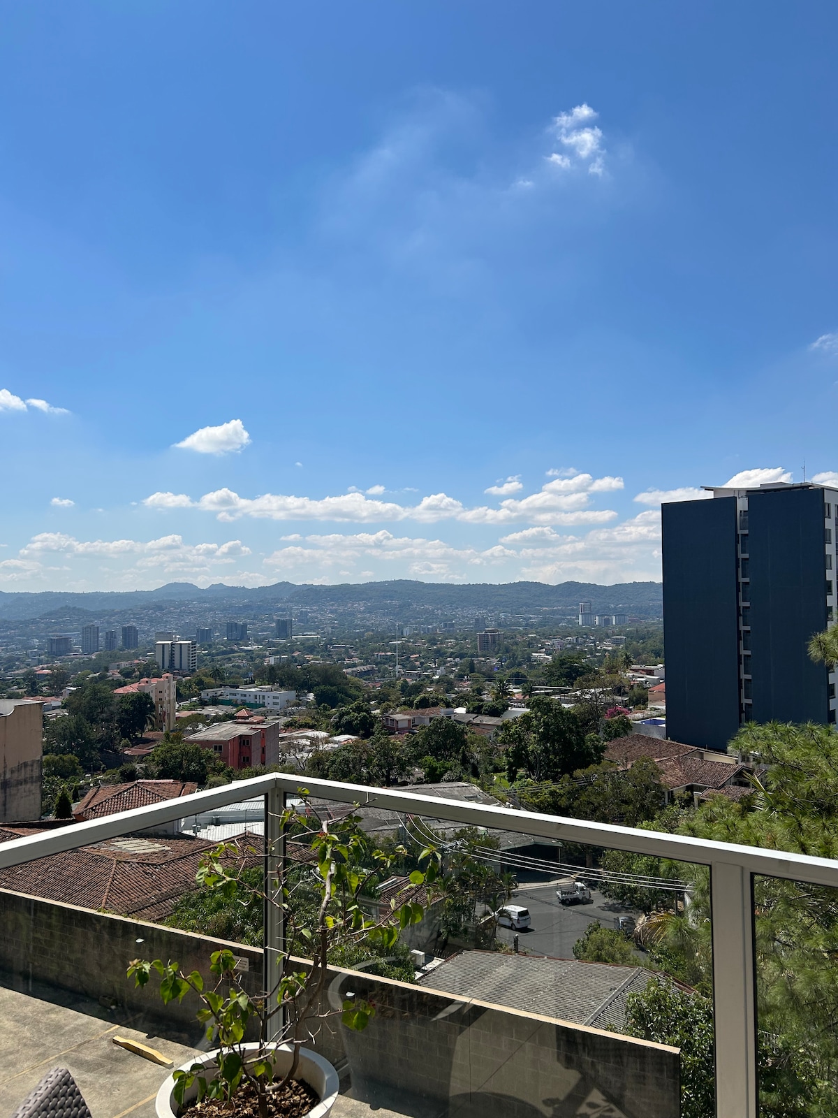 A scenic terrace view features rolling hills and a cityscape under a clear blue sky with fluffy white clouds. The railing offers a transparent barrier, adding to the openness of the space. A small plant in a pot enhances the connection to nature.