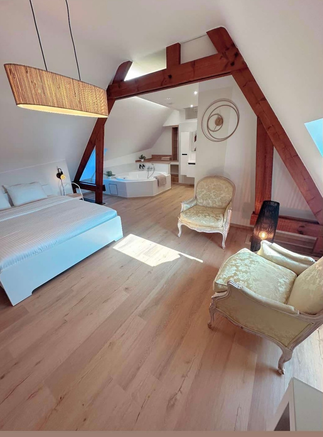 A spacious loft bedroom is depicted, featuring a large bed with soft linens. Elegant armchairs are placed nearby, surrounded by wooden beams that add structure. A bright, inviting atmosphere is created by natural light streaming in through a skylight.