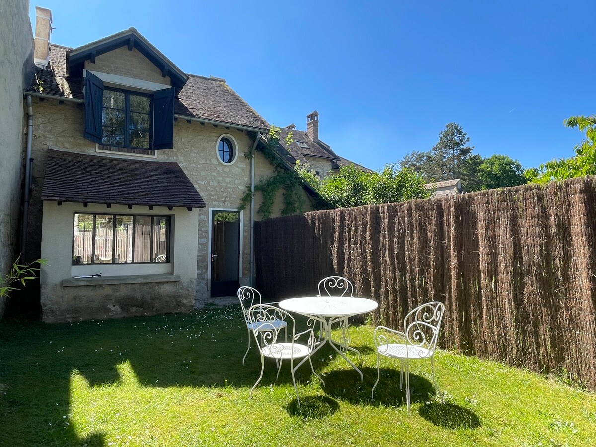 A private outdoor space features a small round dining table surrounded by four white wrought-iron chairs. The lush green lawn is framed by a natural bamboo fence, and the charming stone house stands under a clear blue sky, enhancing the serene setting.
