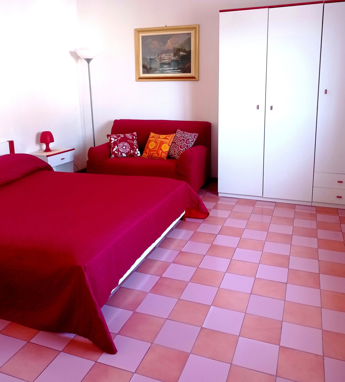 A cozy bedroom features a spacious bed adorned with a burgundy coverlet. A bright red sofa with decorative cushions is positioned alongside, complementing the warm color palette. The room includes a wardrobe and a bedside table, while large tiles in shades of pink provide visual interest.