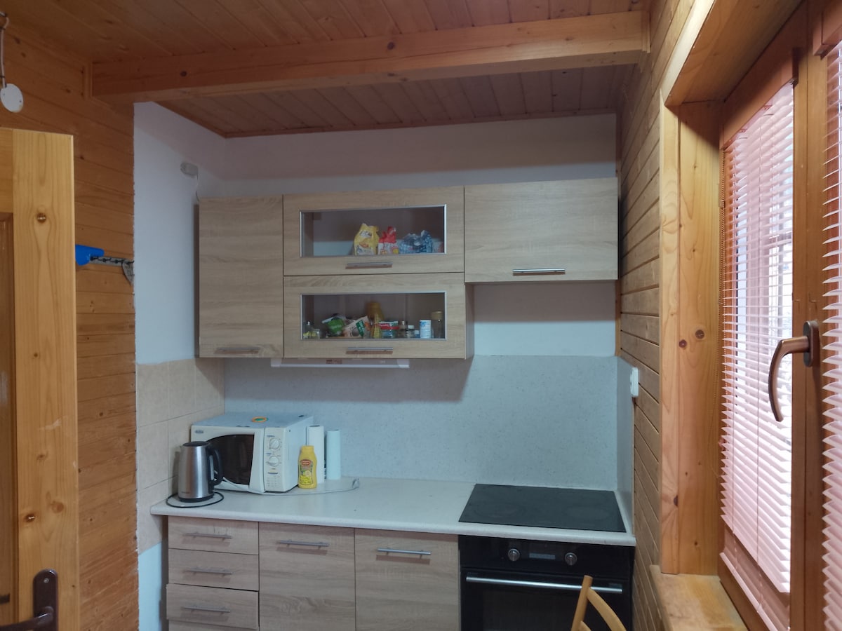 A well-organized kitchen area is shown, featuring wooden cabinetry and a light countertop. Appliances include a kettle, microwave, and an oven. Natural light is allowed in through the window adorned with wooden blinds.