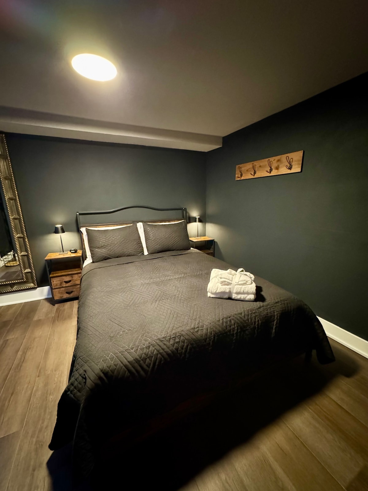 A cozy sleeping area features a spacious bed with a textured dark quilt and decorative pillows. Soft bedside lamps provide gentle illumination, while a unique wooden piece with wall hooks adds a touch of charm. The room is framed by modern wood flooring.