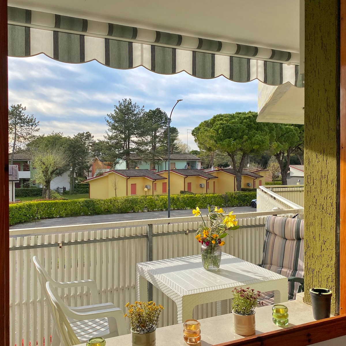 A sunlit terrace is presented, featuring a white table with a small vase of yellow flowers at its center. Two striped chairs are positioned beside the table. A green and white striped awning provides shade, while a quiet street lined with trees and pastel-colored houses can be seen in the background.