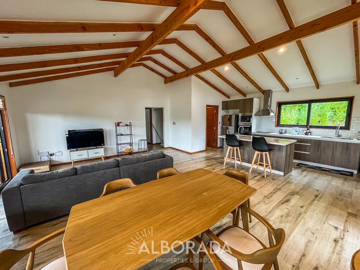 An open living area features a dining table positioned near a modern kitchen and a comfortable seating arrangement. Large windows invite natural light, highlighting wooden beams across the ceiling. A television is mounted on the wall opposite the seating area, creating a welcoming gathering space.
