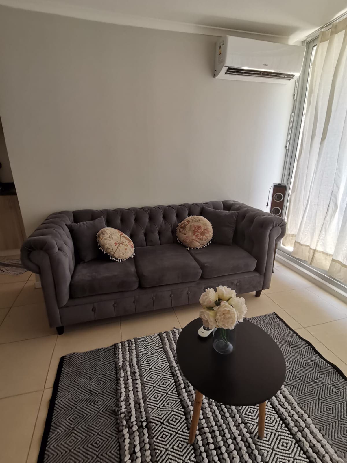 A cozy living area features a tufted gray sofa with decorative cushions, positioned beside a small round table adorned with a vase of fresh flowers. Light streams in through sheer curtains, enhancing the ambient atmosphere, while a patterned rug adds texture to the tiled floor.