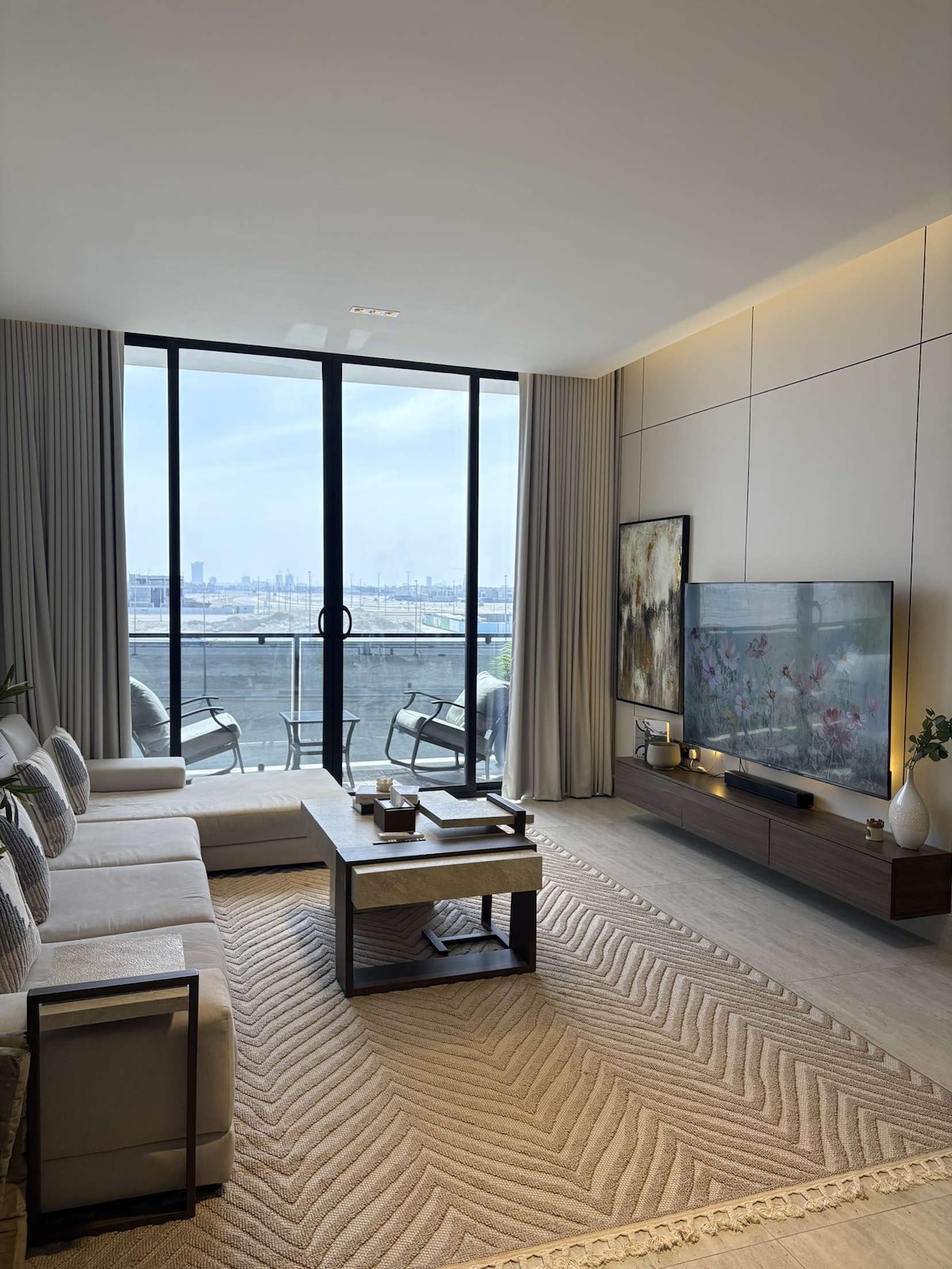 A modern living area is presented with ample natural light from floor-to-ceiling glass doors that open to a balcony. A comfortable seating arrangement is complemented by a stylish coffee table and a large television mounted on the wall. Decorative elements include a soft area rug and contemporary artwork.