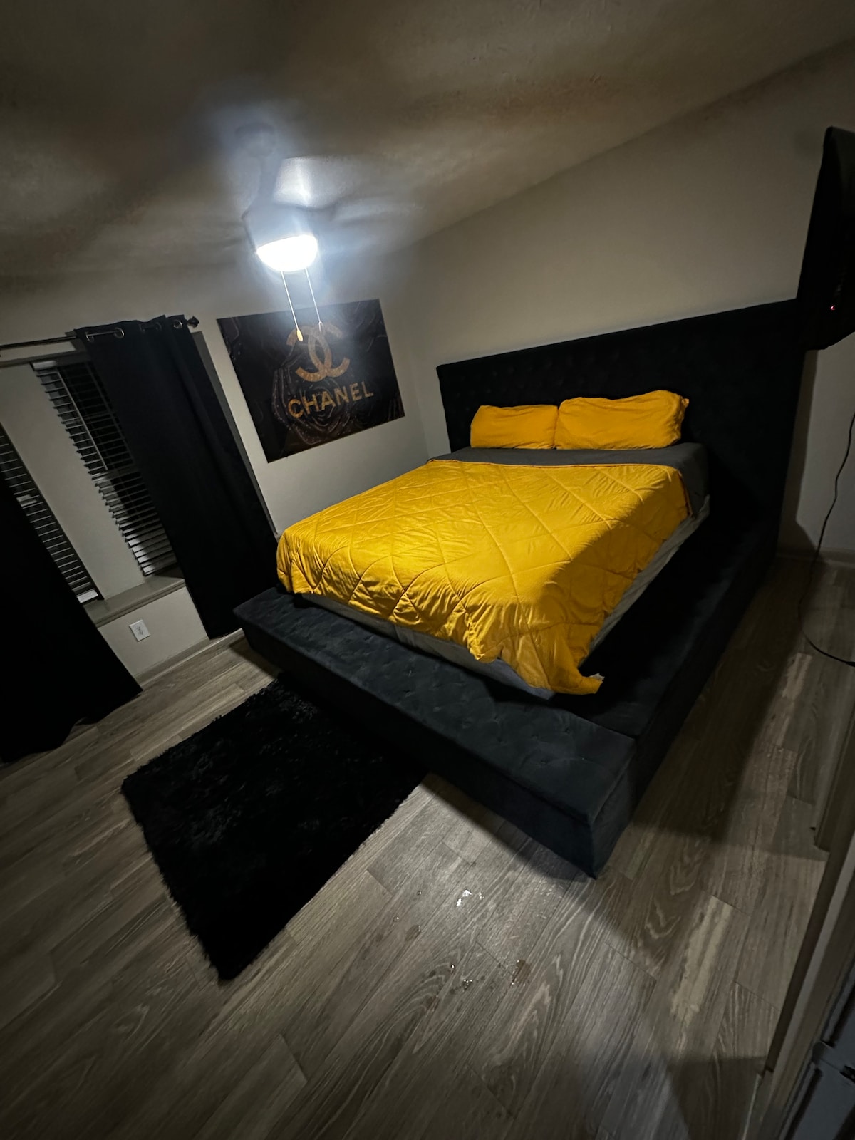 A contemporary bedroom showcases a spacious bed dressed in a bright yellow comforter. A black upholstered frame offers a modern aesthetic. Natural light filters through the window adorned with black curtains, while a stylish area rug adds texture to the wood-like flooring.