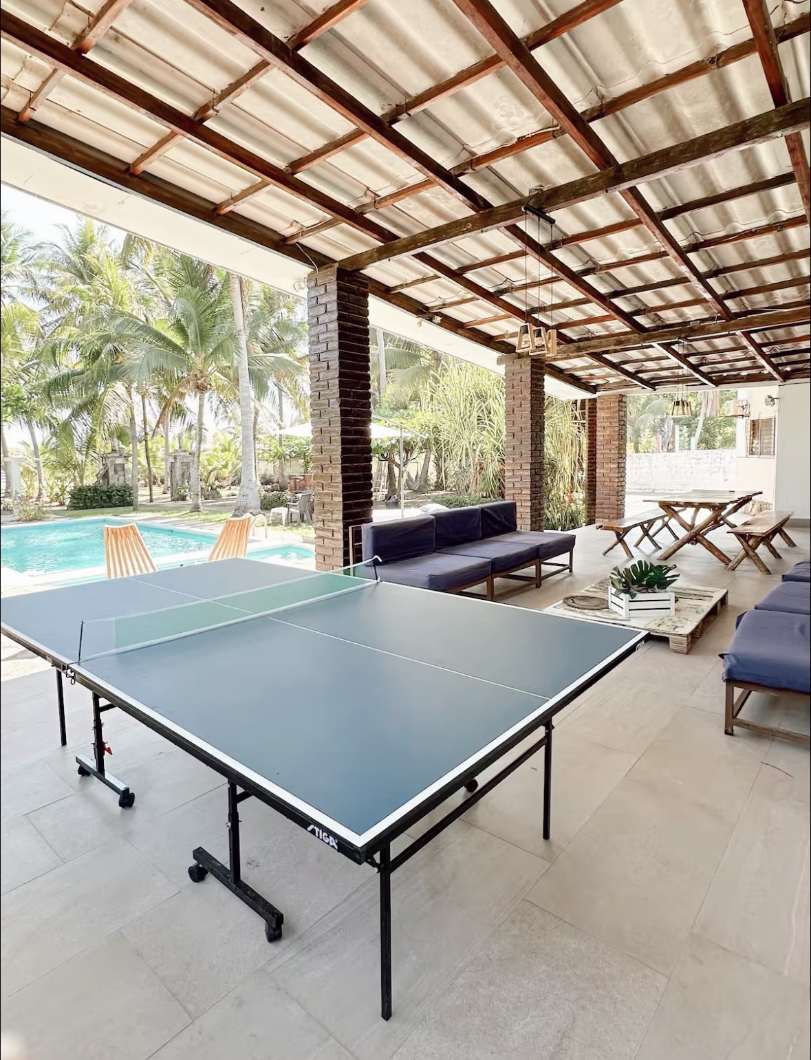 An outdoor seating area features a table tennis setup on a spacious patio. Comfortable seating, including couches and a dining table, surrounds the area, while palm trees and a pool are visible in the background, creating a relaxed environment for guests.
