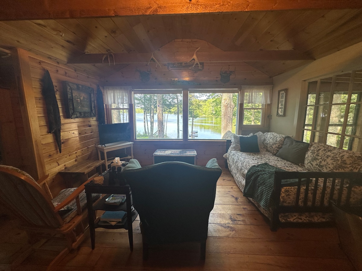A warm living space features a floral-patterned couch, oversized chairs, and a wooden coffee table. Large windows provide views of the lake and surrounding trees. Natural wood finishes and soft lighting create a welcoming atmosphere, complemented by a rustic rocking chair in one corner.