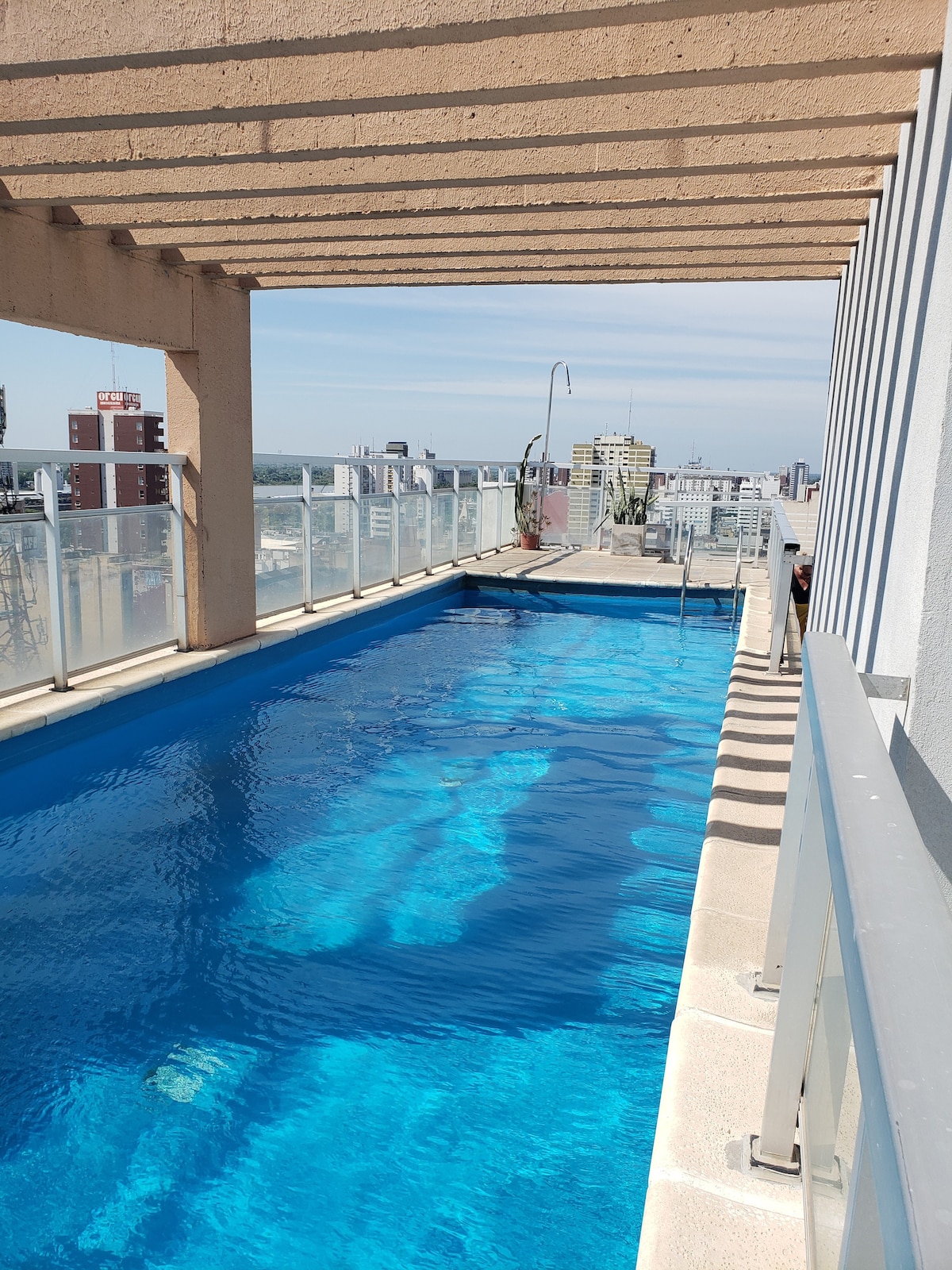 An outdoor pool is situated on a terrace, characterized by clear blue water and a panoramic view of the skyline. Sunlight reflects off the surface of the water, and a swimming lane is visible adjacent to a railing. The surrounding area features a modern architectural design.