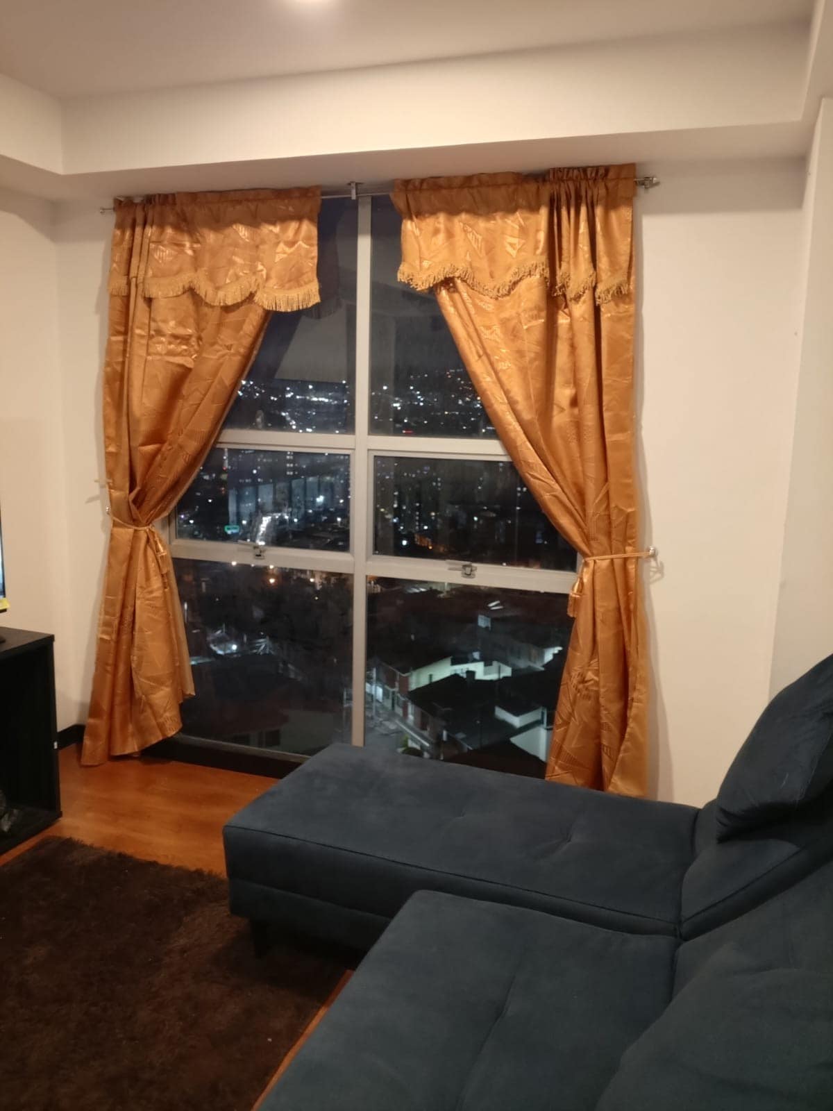 A comfortable seating area features a dark blue sectional sofa, positioned near large windows adorned with golden curtains. The curtains frame a panoramic view of the city lights twinkling in the night sky, providing a sense of urban tranquility.