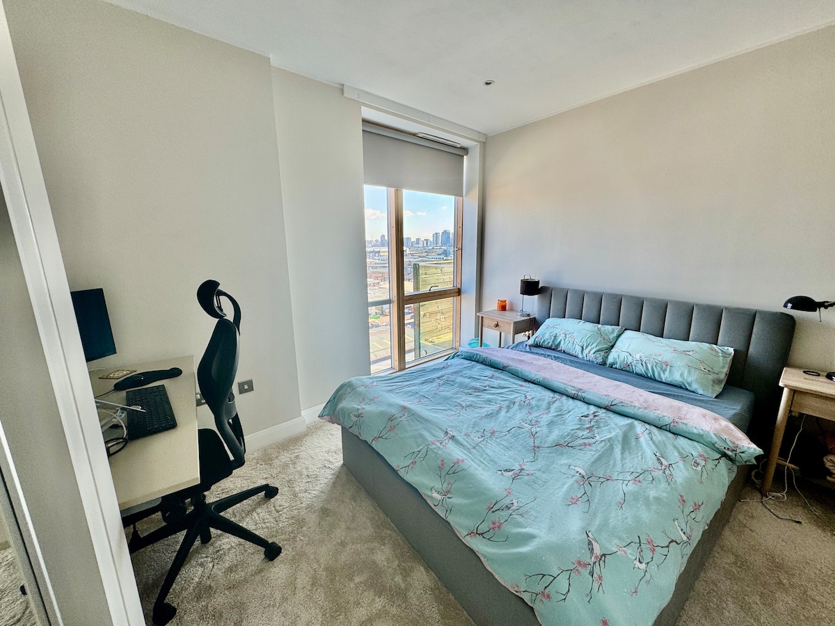 The bedroom features a spacious bed with a light blue duvet set against a neutral wall. A desk with an ergonomic chair is positioned nearby, and a large window provides a view of the cityscape. Natural light brightens the soft beige carpet.