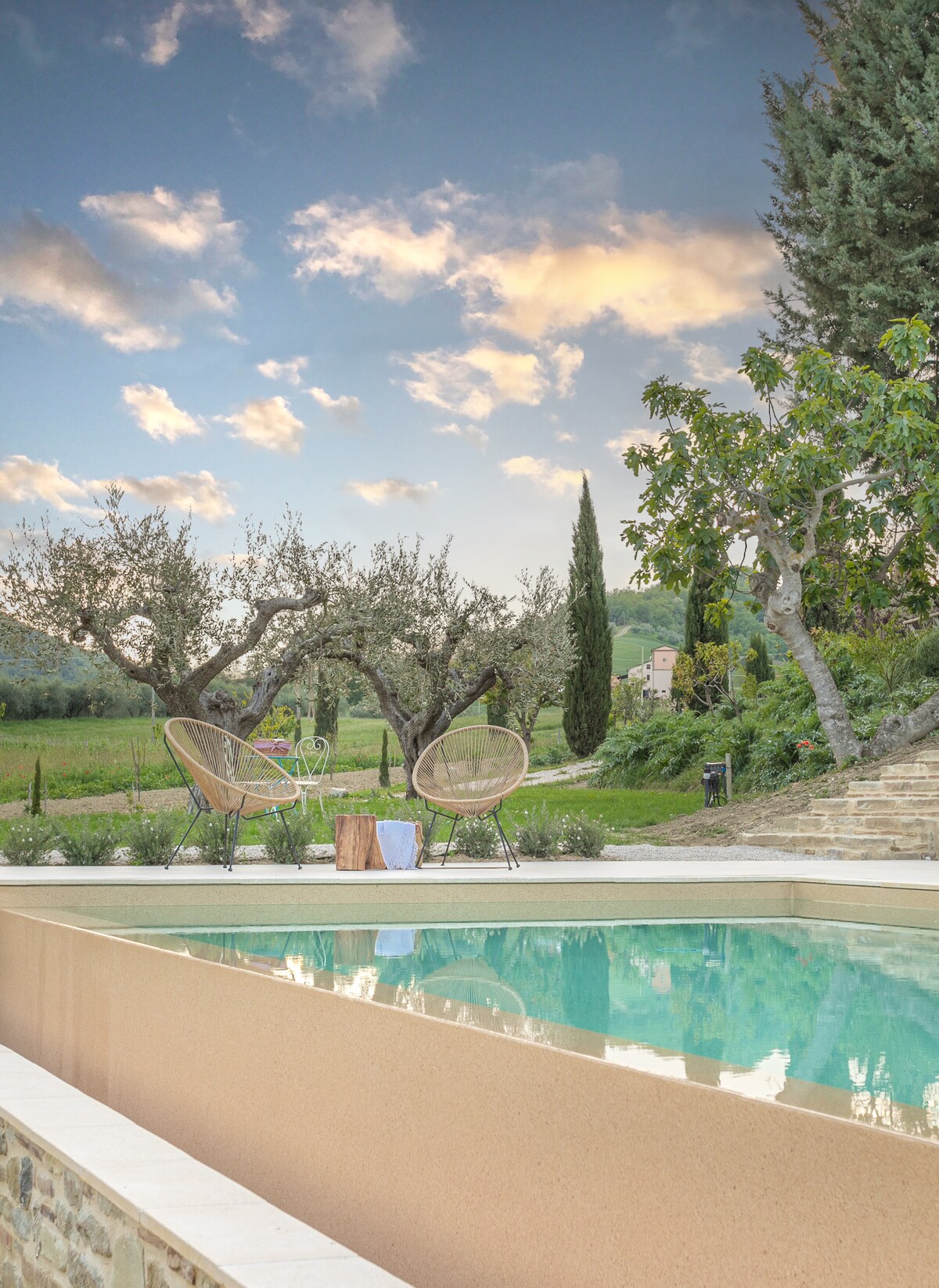 A tranquil pool is set against a scenic backdrop of olive trees and rolling hills. Two comfortable chairs are situated beside the pool, accompanied by a small wooden table. The sky features soft clouds, adding a serene ambiance to the outdoor space.