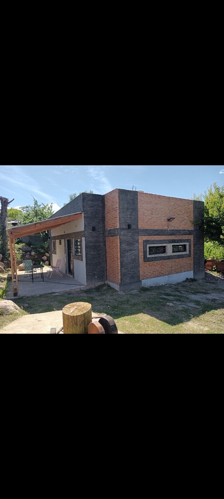 A modern structure features a mix of brick and stone elements, surrounded by greenery. A covered porch is visible, offering seating that creates a welcoming space to enjoy the outdoors. The bright sky highlights the tranquility of the surrounding area.